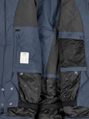 Nahaufnahme einer blauen Jacke mit Reißverschlusstaschen, Innentaschen aus schwarzem Netzstoff und einem weißen Etikett. Das Innenfutter ist schwarz und gesteppt. Sichtbar sind Knopfdetails.