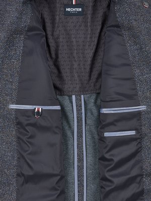 Nahaufnahme der Innenseite eines dunklen Herrenjackets. Der Futterstoff ist dunkelgrau mit einem Muster aus kleinen "H"-Zeichen. Auf der linken Seite befindet sich eine Innentasche mit Knopf und kleinem Emblem, die rechte hat zwei. Über dem Futter steht "HECHTER PARIS".