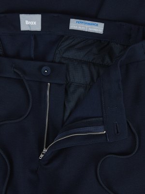 Close-up of navy blue pants. The pants have a zipper, button closure, and drawstring. A tag at the waistband reads "Brax." Another tag reads "Performance, Style Blue, Tapered Fit." The pants are partially open, showcasing interior fabric.