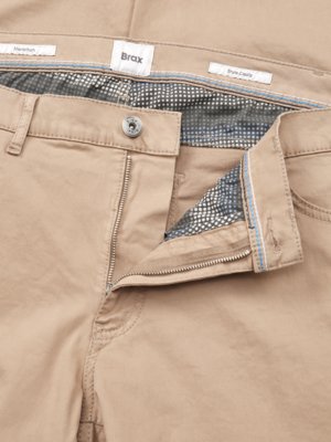 Close-up of beige pants with an unzipped fly. A silver button and a zipper are visible, along with the brand label "Brax." The pants' interior lining has a patterned, mesh-like appearance. Labels read "Marchen" and "Style Coole."