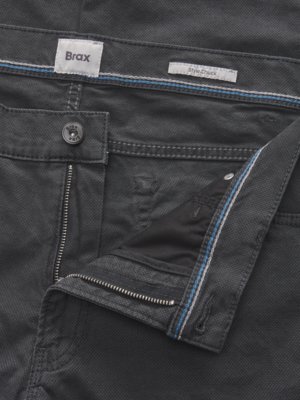 Nahaufnahme einer geöffneten grauen Hose. Deutlich sichtbar sind der Reißverschluss, ein Knopf mit dem Brax-Logo, das Etikett "Brax" und ein weiteres Etikett "Style Chuck". Der Stoff hat ein strukturiertes Muster und blaue Ziernähte.