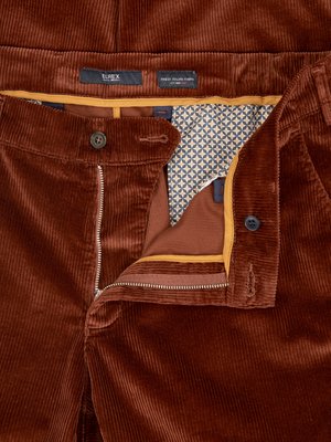 Nahaufnahme einer geöffneten braunen Kordhose. Die Hose ist aus samtartigem Material und hat eine offene Front mit Reißverschluss. Der Bund ist offen, darunter ist ein gemustertes Futter sichtbar. Auf dem Bund sind zwei Etiketten angebracht.