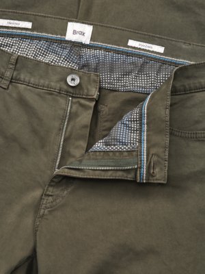 Nahaufnahme einer geöffneten Hose in Olivgrün. Der Reißverschluss ist halb geöffnet, darüber ein Knopf. Innenfutter mit kleinem Muster. Über der Knopfleiste ist ein Label der Marke Brax zu sehen.