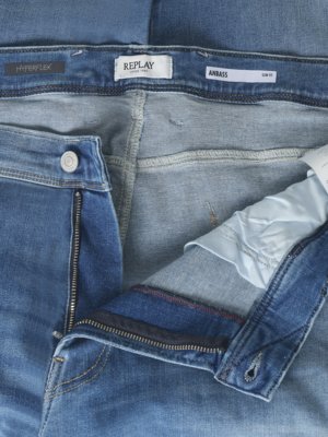 Detailaufnahme einer hellblauen Jeanshose. Die Hose ist geöffnet, die Knöpfe und der Reißverschluss sind sichtbar. Oben ist ein Markenetikett "REPLAY" zu sehen, daneben "ANBASS Slim Fit".