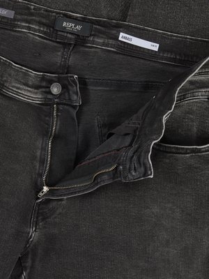Nahaufnahme einer Jeans in Used-Optik. Der Reißverschluss ist offen. Ein Knopf und die Markenbezeichnung „REPLAY“ sind sichtbar, daneben der Schriftzug „ANBASS SLIM FIT“. Die Hose ist dunkelgrau.