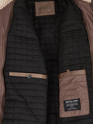 Nahaufnahme des Innenfutters einer braunen Jacke. Innenfutter aus gestepptem schwarzem Stoff. Aufgenähte braune Lederapplikationen mit Knöpfen und Taschen. Ein Etikett mit Text "VEGETABLE TANNED" ist sichtbar.