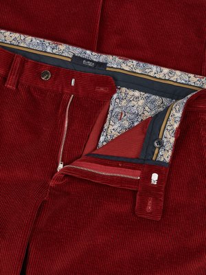 Naheinstellung einer offenen roten Cordhose. Der Reißverschluss ist halb geöffnet, ein Knopf ist geknöpft. Das Innenfutter zeigt ein Muster in Blau- und Beigetönen, ein EUREX-Etikett ist sichtbar.