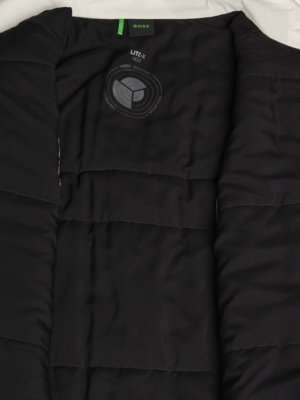 Innenansicht einer schwarzen Weste mit weißem Futter. Oben steht BOSS in Grün. Unten rechts befindet sich ein Kreis mit der Aufschrift LITE-X Vest, Water Resistant. Die Weste ist offen, die Steppung der Innenseite ist sichtbar.