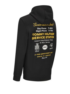 Schwarzer Kapuzenpullover mit gelbem Aufdruck auf dem Rücken. Der Aufdruck zeigt den Text "Service with a Smile", Telefonnummern und "Tommy Hilfiger Service Station" mit Zusatzleistungen.
