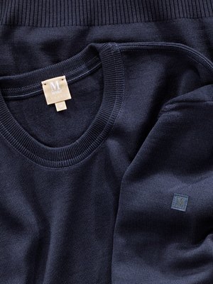 Nahaufnahme von zwei dunkelblauen Strickpullovern. Einer der Pullover hat einen runden Halsausschnitt und ein Etikett mit der Aufschrift "ML". Auf dem anderen Pullover ist ein kleines quadratisches Logo sichtbar.