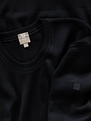 Nahaufnahme eines schwarzen Sweatshirts. Der Rundhalsausschnitt und das Etikett mit dem Logo "M MARQZ" sind deutlich sichtbar. Am Ärmel ist ein eckiger Aufnäher mit Logo. Der Stoff ist fein strukturiert.