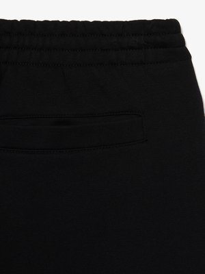 Nahaufnahme von schwarzer Hose, die aus grobem Stoff zu bestehen scheint. Der obere Bereich zeigt einen elastischen Bund mit horizontalen Nähten. Ein rechteckiger Hosentaschenausschnitt ist deutlich zu sehen. Der Stoff hat eine glatte Oberfläche.