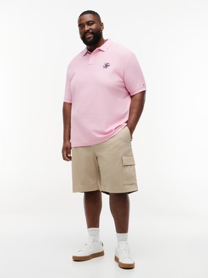 Ein Mann in einem rosafarbenen Poloshirt und einer beige Cargohose steht vor einem weißen Hintergrund.