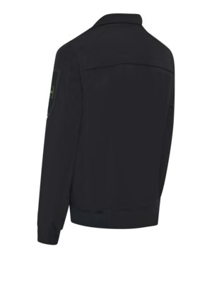 Rückansicht einer schwarzen Jacke mit grau-grünem Ärmelabzeichen und einem durchgehenden Reißverschluss.
