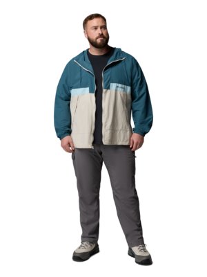 A man stands wearing a color-blocked hooded jacket in teal, light blue, and beige, with grey cargo pants and hiking boots.