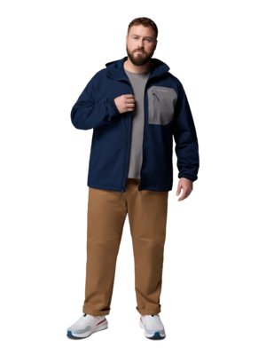 Ein Mann mit Bart steht frontal und trägt eine dunkelblaue Jacke mit grauer Brusttasche, ein graues T-Shirt, Khakihosen und weiße Sportschuhe.