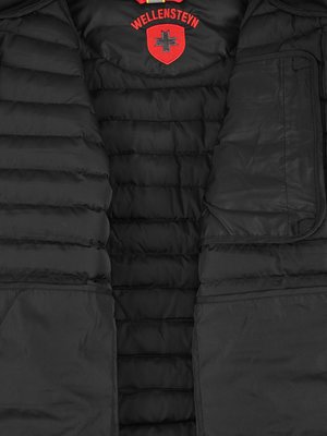 Nahaufnahme des Innenfutters einer schwarzen Steppjacke. Im oberen Bereich befindet sich ein rotes Logo-Emblem mit dem Schriftzug "WELLENSTEYN". Die Jacke hat gepolsterte Nähte und Innentaschen.