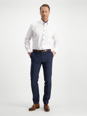 Ein Mann in einem weißen Hemd, einer blauen Hose und braunen Schuhen posiert vor hellem Hintergrund. Er hat die Hände in den Hosentaschen und trägt einen braunen Gürtel. Sein Hemd ist aufgeknöpft.