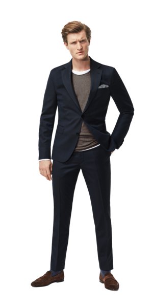 A man wearing a navy blue suit over a brown sweater and white undershirt stands facing the viewer. He has short, light brown hair and is wearing brown loafers and blue socks. He has a pocket square.