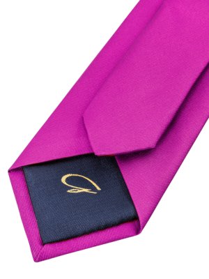 Nahaufnahme einer Krawatte. Der Großteil der Krawatte ist aus magenta-farbenem Stoff. Auf der Innenseite der Krawatte befindet sich ein dunkelblauer Stoff mit einem goldenen Logo.