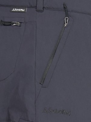 Detaillierte Nahaufnahme einer dunklen Hose mit Reißverschlusstaschen. Ein seitlicher Reißverschluss und ein Logo "Schöffel" sind sichtbar. Die Hose hat eine glatte Oberfläche und ist vermutlich aus einem technischen Material gefertigt.