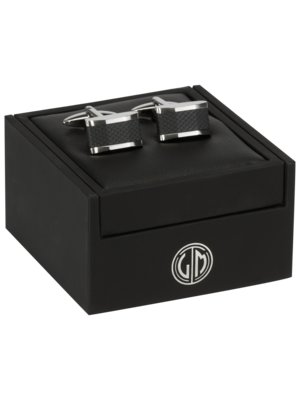 Ein Paar Manschettenknöpfe liegt in einer schwarzen, rechteckigen Box. Die Manschettenknöpfe sind rechteckig mit schwarzem Muster und silberfarbenen Rändern. Auf der Box ist ein Logo.