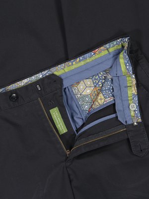 Nahaufnahme einer schwarzen Hose. Der Bund ist mit einem farbigen Muster versehen, und das Innenfutter ist blau mit einem farbigen Dekor. Reißverschluss und Knopfverschluss sind sichtbar. Ein grünes Etikett ist angenäht.