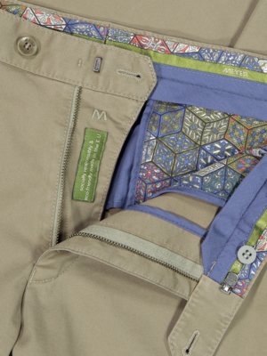 Naheinstellung einer beigefarbenen Hose, teilweise geöffnet. Details wie Reißverschluss, Knopf, Innenfutter mit Muster und ein grünes Etikett mit Aufschrift "sozial verantwortlich & umweltfreundlich". "MEYER" Label.