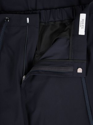 Nahaufnahme der geöffneten dunklen Hose. Ein Reißverschluss und ein Knopf sind sichtbar. Am Bund befindet sich ein Gummiband und ein Logo-Etikett. Eine Kordel ist links zu sehen. Die Hose ist aus Stoff.