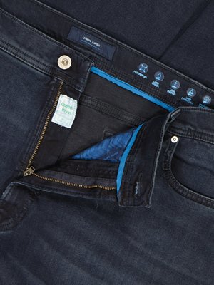 Nahaufnahme einer Jeans in dunkler Farbe, geöffnetem Reißverschluss und blauem Futter. Auf der Jeans steht "Pierre Cardin" und "Green Rivet". Um den Bund sind Symbole abgebildet.