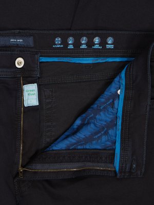 Close-up of black jeans, unbuttoned and unzipped. The jeans feature a tag that says "Green Rivet." Blue accents are visible along the waistband and inside the fly. Above, various icons indicate features like "Organic Cotton." The brand is Pierre Cardin.
