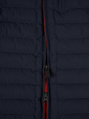 Nahaufnahme einer dunkelblauen Steppjacke mit schwarzem Reißverschluss. Der Reißverschluss verläuft mittig, mit roter Einfassung. Die Jacke hat horizontale Steppnähte. Der Reißverschluss ist geschlossen.