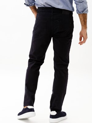 A man from the back is shown wearing dark blue pants, light blue shirt, and dark blue shoes with white soles. His right hand is in his pocket. He is wearing a woven belt. The background is white.
