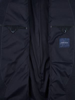 Nahaufnahme des Innenteils einer dunkelblauen Jacke. Zwei kleine Taschen, eine mit einem runden Knopf. Ein blaues Etikett mit der Aufschrift "Jersey". Die Innenseite der Jacke ist aus glänzendem Stoff.