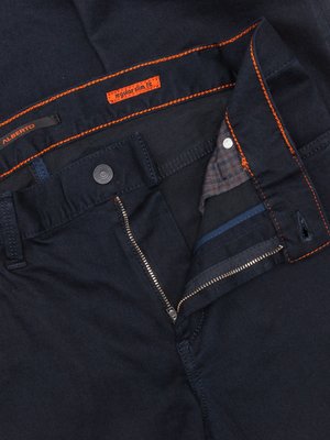 Nahaufnahme einer dunklen Jeanshose, teilweise aufgeknöpft und geöffnet. Der Reißverschluss ist geöffnet. Orangefarbene Nähte sind zu sehen, ebenso ein Etikett mit "regular slim fit". Auf einem Etikett steht "ALBERTO". Der Hosenknopf ist aus Metall.