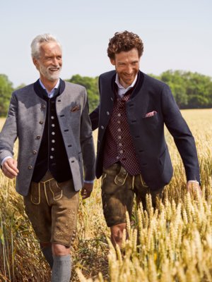 Zwei Männer spazieren durch ein Getreidefeld. Beide tragen Trachtenjacken, Westen und Lederhosen. Der ältere Mann links hat graue Haare, der jüngere rechts hat Locken. Sie lächeln beide.