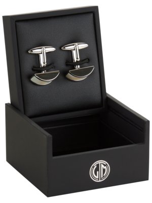 Schwarze Manschettenknöpfe liegen in einer geöffneten schwarzen Geschenkbox. Die Manschettenknöpfe haben eine silberne Oberfläche und eine schwarze, halbkreisförmige Verzierung. Auf der Box ist ein silbernes Logo.