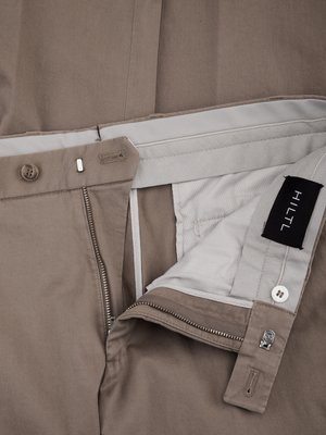 Nahaufnahme einer geöffneten, beigen Hose, Detailansicht von Knopfleiste, Reißverschluss und Hosenbund. Ein schwarzes Etikett mit "HILTL" ist innen sichtbar. Knöpfe und metallene Details sind zu sehen.