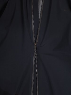 Nahaufnahme einer schwarzen Jacke mit Reißverschluss in der Mitte. Der Reißverschluss ist geschlossen. Der Stoff der Jacke ist dunkelblau und leicht glänzend. Der Hintergrund ist hellgrau.