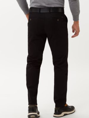 Ein Mann von hinten in schwarzer Hose, grauer Jacke und schwarzen Schuhen. Er trägt einen schwarzen Gürtel und hat die Hände in den Hosentaschen. Der Hintergrund ist hell.