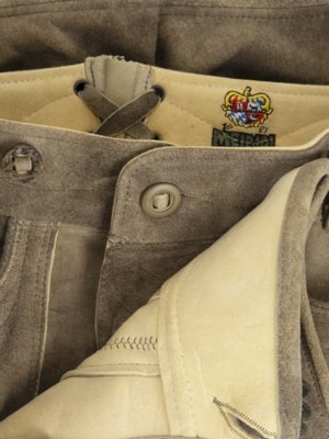 Nahaufnahme einer traditionellen Lederhose in Braun. Die Hose hat Knöpfe, eine Schnürung am Bund und ein Label mit Wappen. Ein Stück des hellen Innenfutters ist sichtbar.