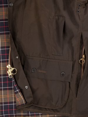 Nahaufnahme einer braunen Wachsjacke. Die Jacke hat eine goldfarbene Reißverschluss, zwei Taschen, Knöpfe und ein Tartan-Innenfutter. Das Barbour-Logo ist auf einer der Taschen deutlich sichtbar.