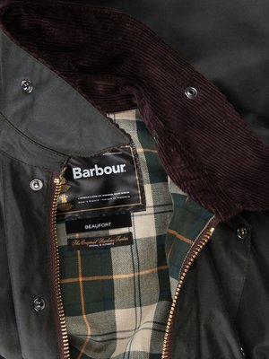 Detaillierte Nahaufnahme einer grünen Barbour-Jacke. Der Kragen besteht aus braunem Cord, darunter ein Innenfutter mit grünem Karomuster. Ein Etikett mit "Barbour" und "Beaufort" ist sichtbar. Die Jacke ist teilweise geöffnet.