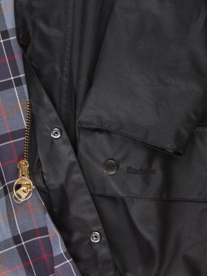 Detailaufnahme einer schwarzen Jacke, teilweise geöffnet. Das Innenfutter zeigt ein blau-graues Karomuster, die Jacke hat eine goldfarbene Reißverschluss und Knöpfe. Auf einer Tasche ist "Barbour" gestickt.