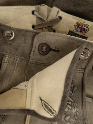 Nahaufnahme einer graubraunen Lederhose. Knöpfe, Details von Nähten und Verzierungen sind sichtbar. Der Bund ist mit Schnürsenkeln versehen. Ein Emblem ist am Bund zu sehen. Das Innenfutter ist hell.