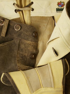 Detailaufnahme von Lederbekleidung, darunter eine braune Lederhose mit Knöpfen, hellfarbene Lederdetails und ein weißes Oberteil mit Emblem und schwarzem Knopf. Verschiedene Texturen.