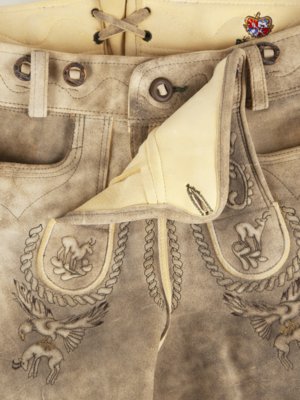 Nahaufnahme einer hellbraunen Lederhose. Der Hosenlatz ist teilweise geöffnet und zeigt einen helleren Stoff. Stickereien mit Tierdarstellungen und Zierkordeln sind zu sehen. Details wie Knöpfe, Hosenbund und Zierschnüre sind erkennbar.