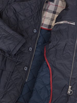 Nahaufnahme einer dunkelblauen, gesteppten Jacke, teilweise geöffnet. Innenfutter mit blauem Rautenmuster und roter Naht. Oberteil mit Tartanmuster und Reißverschluss. Knöpfe und Innentasche sichtbar.