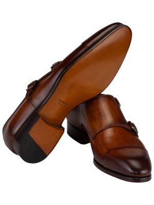 Zwei braune Lederschuhe mit Schnallenverschlüssen. Einer der Schuhe ist auf dem Kopf gedreht, so dass die braune Sohle zu sehen ist. Der andere Schuh steht aufrecht. Die Schuhe haben abgerundete Kappen und sind auf weißem Hintergrund fotografiert.
