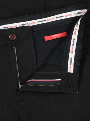 Nahe Aufnahme einer schwarzen Hose. Der Hosenbund ist geöffnet, ein brauner Knopf ist sichtbar. Am Bund ist das Logo "TOMMY JEANS" zu sehen. Ein rotes Etikett mit der Aufschrift "SCANTON" ist ebenfalls sichtbar.